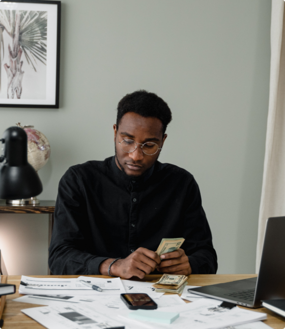 A man wearing glasses sits at his desk in front of a laptop holding money and looking and financial documents