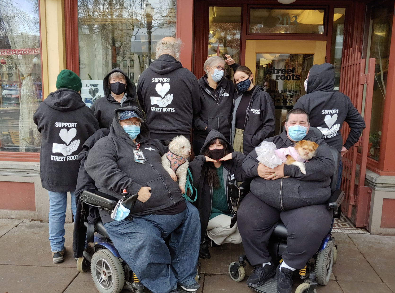 Street Roots team, in hoodies saying "support street roots," wearing masks, in front of a store front.