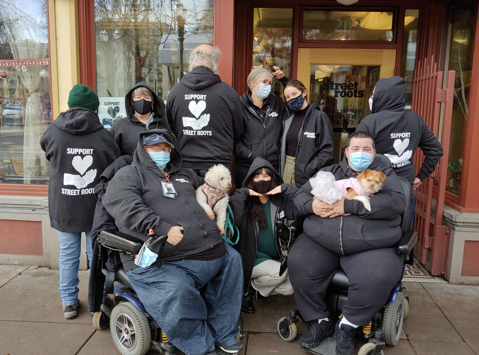 A group of people in masks pose together in Street Roots hoodies in front of a storefront