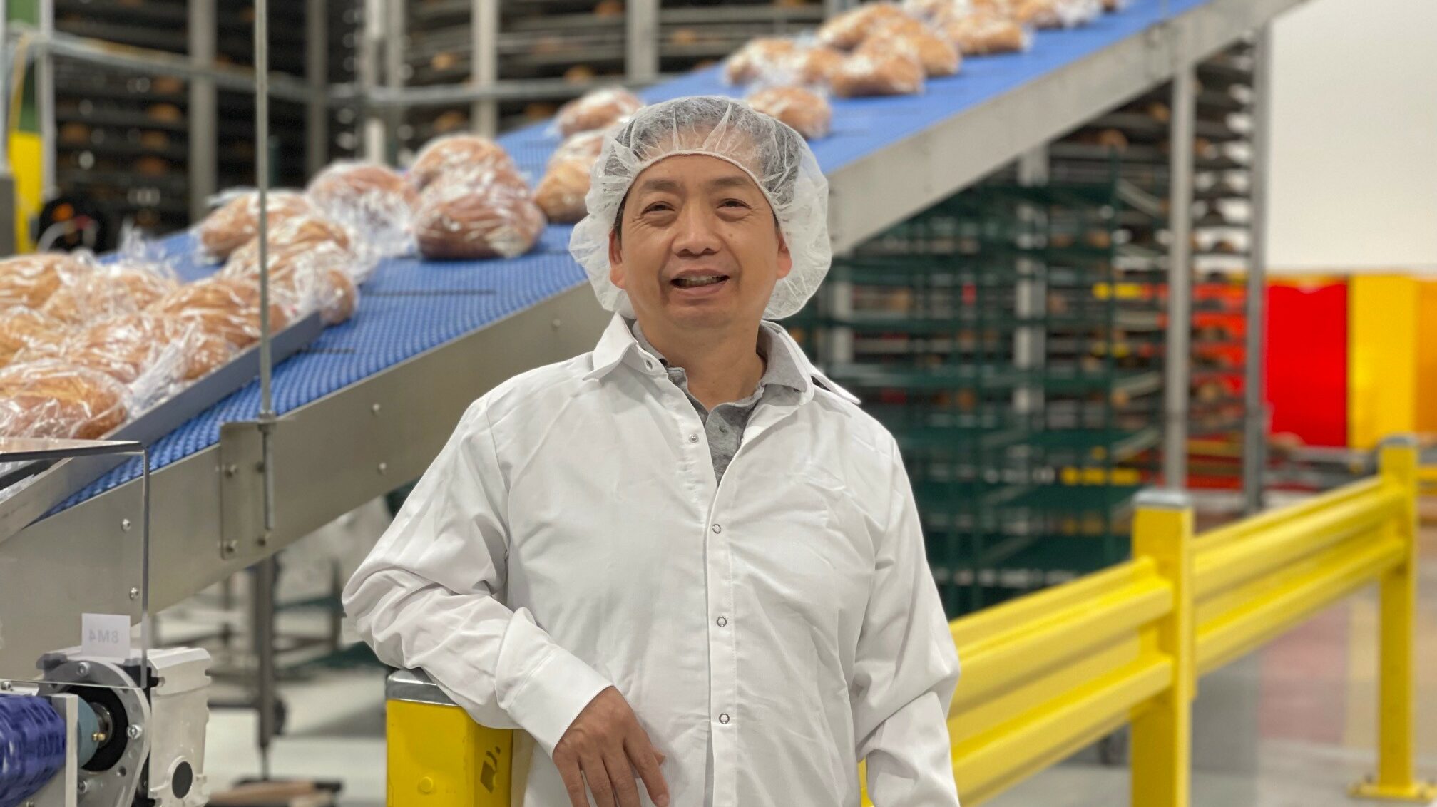 United Bakery owner and founder Dinh Nguy stands in front of a sourdough bread conveyor belt in a factory.