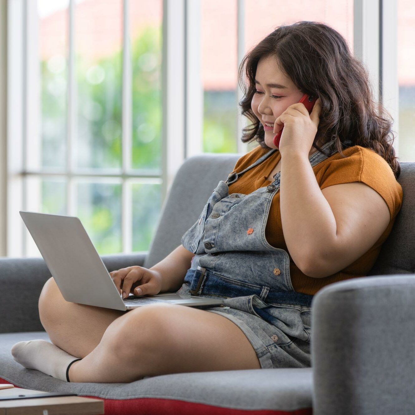 Woman wearing overalls sits on a couch while talking on the phone and using her computer