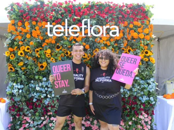 Two people stand in front of a wall of flowers with signs that read "Queer to Stay" and "Queer Se Queda," smiling at the camera.