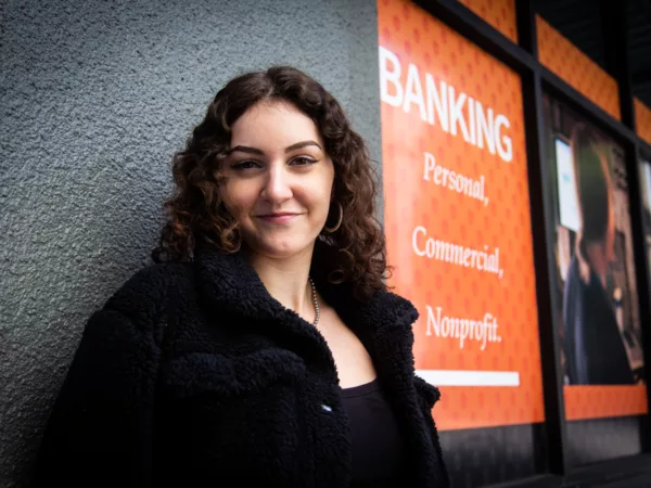 Sierra Carroll has brown curly hair and wears a black jacket and top. She stands in front of Beneficial State Bank's Pearl Branch in Portland, Oregon, smiling at the camera