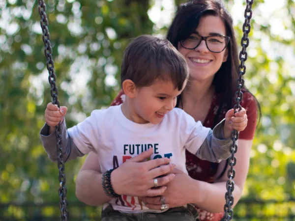 Woman with brown hair and glasses hugging a kid on a swing. Both are smiling.