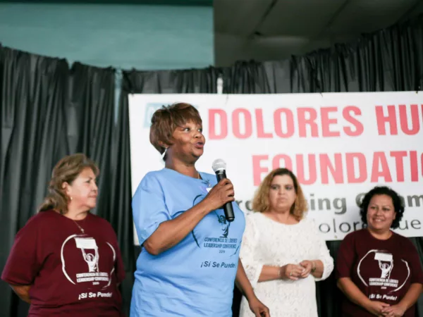 Four people stand on a stage in front of a Dolores Huerta Foundation banner. One person in a blue shirt stands in the foreground holding a microphone.