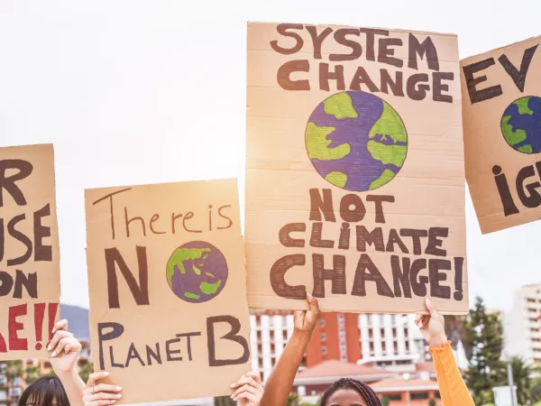 Four pairs of arms hold up signs calling on leaders to address climate change. The signs say "our house is on fire," "There is no planet B," "system change not climate change." and "evidence over ignorance" ("evidence" and "ignorance" are slightly cut off)