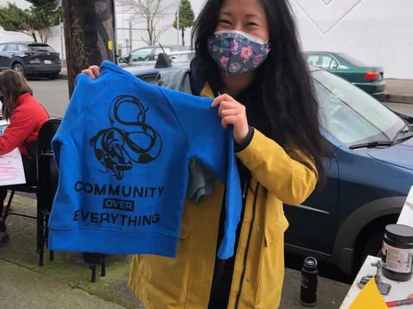 Asian woman wearing a mask and a yellow jacket holds up a dark blue sweatshirt that says "Community Over Everything"