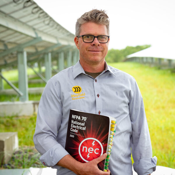 Ryan Mayfield, the Founder and Owner of Mayfield Renewables, holds a National Electrical Code textbook while standing in a large solar farm