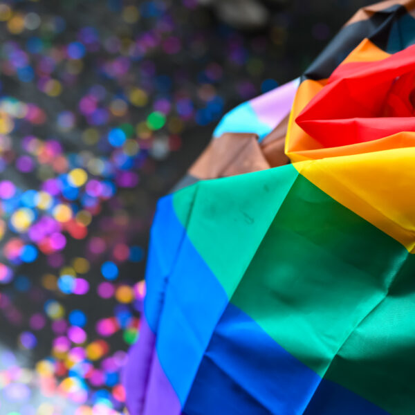 A person draped in a rainbow flag stands amidst colorful confetti.